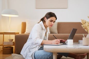 Side view full length young focused female in casual clothes browsing modern laptop and writing down notes while sitting on floor in cozy modern apartment